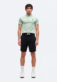 Homme portant un polo vert clair, un short noir avec ceinture, des chaussettes blanches et des baskets blanches, debout sur fond blanc.