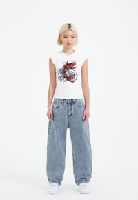 White cotton t-shirt with a red fish graphic and waves, paired with loose blue denim jeans. Model stands with hands at sides.