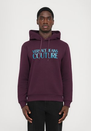 Ung mann iført en mørkerød hettegenser med teksten "Versace Jeans Couture" i blått, stående mot en lys ensfarget bakgrunn.