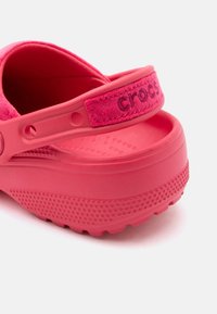 Des sabots Crocs rose vif affichant une semelle texturée, une sangle de talon ajustable avec un patch en tissu et un lit de pied embossé pour le confort et l'adhérence.