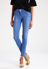 Jeans ajustados de mezclilla en color azul, con un diseño clásico de cinco bolsillos y un dobladillo deshilachado, combinados con zapatos de plataforma dorados.