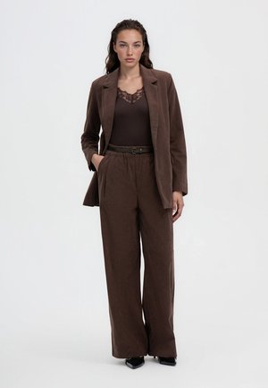 Donna in piedi che indossa un blazer in velluto a coste marrone e pantaloni a gamba larga, top marrone scuro con pizzo e scarpe nere a punta su sfondo bianco.