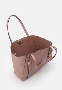 Sac tote Guess rose poudré avec deux bretelles, poche zippée, fermeture à pression magnétique et détail de tag de marque sur fond blanc.