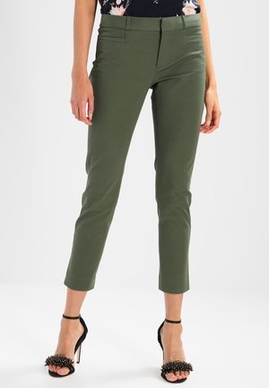 Frau trägt schmal geschnittene olivgrüne Knöchelhosen und schwarze Sandalen mit hohen Absätzen und spitzenbesetzten Riemen, stehend vor einem schlichten Hintergrund.