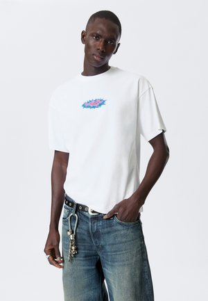 Witte katoenen t-shirt met korte mouwen, voorzien van een multicolore grafisch ontwerp in het midden. Het model draagt een blauwe spijkerbroek met een kettingriem.