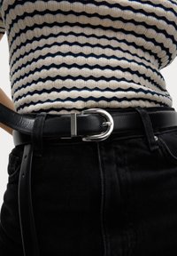 Zwarte leren riem met een zilveren metalen gesp, gedragen over zwarte denim. Gepatternde gebreide top met witte en marineblauwe horizontale strepen.