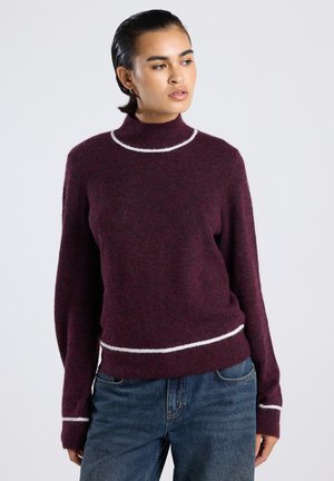 Woman with slicked-back hair wearing a maroon sweater with white trim around the neck, cuffs, and hem, paired with blue jeans.