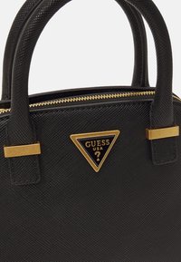 Sac à main noir texturé avec doubles poignées, fermeture éclair dorée et plaque triangulaire du logo Guess USA à l'avant.