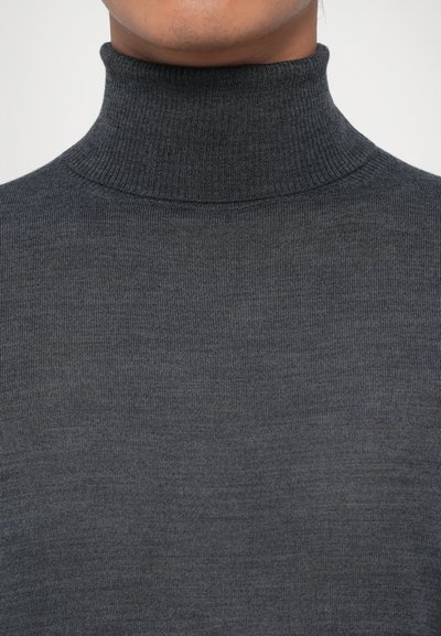 Person, der einen dunkelgrauen, gerippten Rollkragenpullover trägt, der bis unter die Nase gekürzt ist, vor einem schlichten hellen Hintergrund.