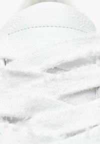 Białe sneakersy z teksturowanymi sznurowadłami i gładką skórzaną cholewką. Sznurówki są puszyste, częściowo zasłaniając język buta oraz szwy.