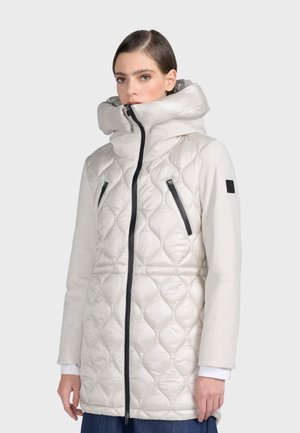 Manteau blanc rembourré avec un col montant, présentant une texture matelassée, des poches zippées noires et des détails zippés noirs contrastants.