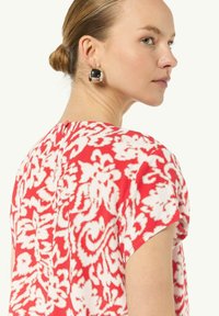 Rot- und weiß geblümte Bluse mit kurzen Ärmeln, die ein Rückenschlüsselloch-Detail aufweist. Das Model trägt runde schwarze und silberne Ohrringe.