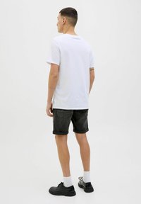 Tricou alb din bumbac, cu mâneci scurte, asortat cu pantaloni scurți negri din denim. Modelul poartă pantofi închiși la culoare și șosete albe, văzut din spate.