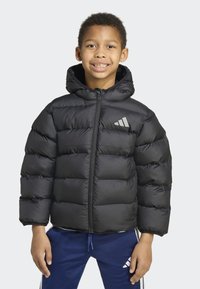 Giacca imbottita nera con cappuccio, caratterizzata da sezioni trapuntate, cerniera frontale e logo grigio sul petto a sinistra. Indossata sopra una camicia bianca.