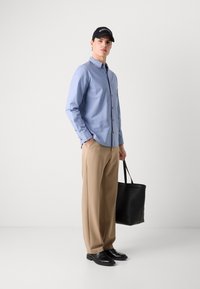 Camisa azul-claro de botões, calças bege, sapatos pretos e uma mala tote preta. A camisa possui um pequeno logo e botões na frente.