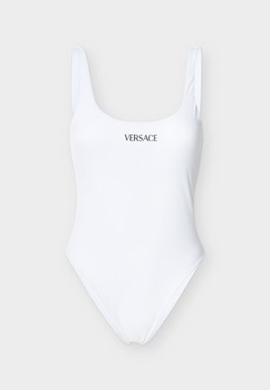 Biały jednoczęściowy strój kąpielowy z szerokimi ramiączkami i czarnym logo "VERSACE" na środku klatki piersiowej na jasnoszarym tle.