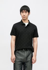 Polo nera con motivo logo testurizzato, maniche corte e colletto nero. Indossata con jeans in denim grigio. Design semplice, senza accenti aggiuntivi.