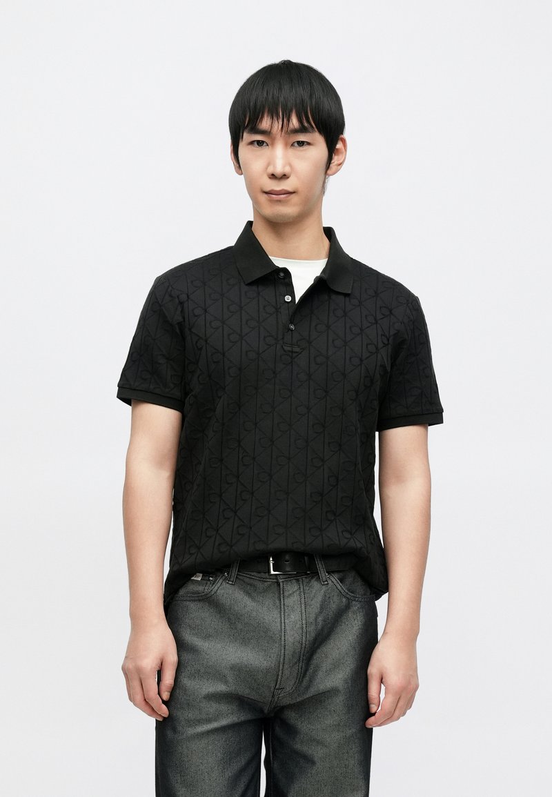 Polo nera con motivo logo testurizzato, maniche corte e colletto nero. Indossata con jeans in denim grigio. Design semplice, senza accenti aggiuntivi.