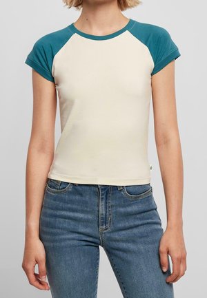 Frau trägt ein beige T-Shirt mit kurzen Ärmeln und türkisfarbenen Raglan-Ärmeln sowie eine blaue, hoch taillierte Jeans und steht vor einem schlichten Hintergrund.