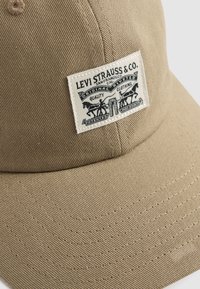 Cappellino da baseball beige realizzato in tessuto testurizzato, con un motivo cucito e un'etichetta rettangolare con grafiche e testo neri.