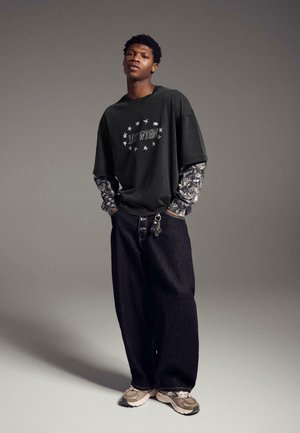 Giovane uomo che indossa una t-shirt nera oversize con stelle, maniche lunghe con motivi sotto, jeans larghi scuri, sneaker beige e portachiavi come accessorio.