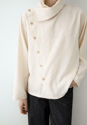 Person trägt ein cremefarbenes Langarmoberteil mit hohem Kragen und diagonalem Knopfverschluss, kombiniert mit dunkler Hose.