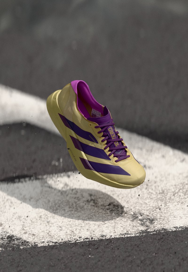 Chaussure de sport jaune avec des accents et des rayures violets, présentant un design épuré, une surface texturée et des lacets contrastants, flottant au-dessus du sol.