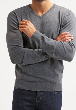 Grauer V-Ausschnitt-Pullover aus weichem Strickstoff. Eng anliegende Ärmel und Saum mit Rippstruktur. Getragen über dunklen Jeans, mit verschränkten Armen.