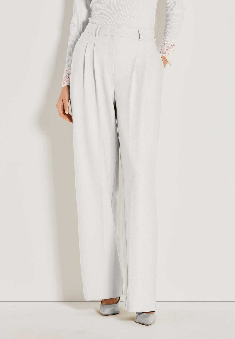 Frau mit hochgeschnittenen, weit geschnittenen weißen Hosen mit Falten, kombiniert mit einem weißen Oberteil und hellgrauen, spitzen Absatzschuhen.