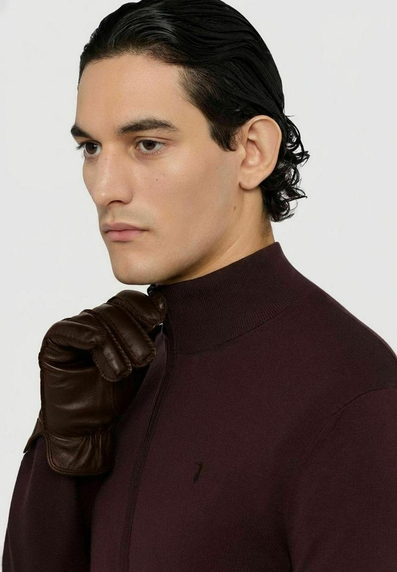 Pull en tricot bordeaux avec un col montant et une fermeture éclair, assorti à des gants en cuir marron. Texture lisse, coupe ajustée, marque discrète.