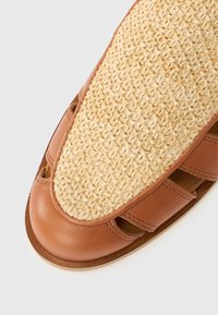Καφέ δερμάτινο σανδάλι με πλεκτό άνω μέρος από άχυρο, διαθέτει σχέδιο με κοψίματα και λεία υφή στο μπροστινό μέρος, συνδυάζοντας φυσικά υλικά και δεξιοτεχνία.
