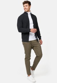 Mørk grå strukturert zip-up genser over en hvit høyhalset genser, parret med olivengrønne slim-fit bukser og hvite sneakers. Renslig, moderne utseende.