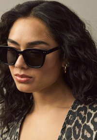 Lunettes de soleil noires rectangulaires avec une finition brillante, dotées de verres foncés et de petits accents en métal. Portées avec un haut à imprimé léopard.