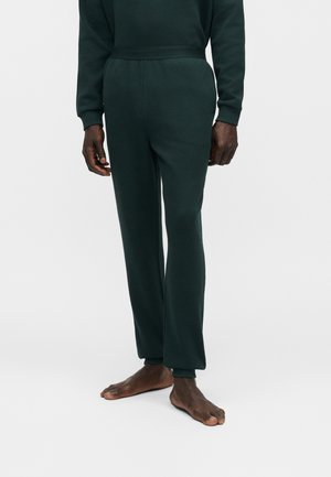 Pantaloni verde scuro con trama e vita elastica indossati da una persona scalza in piedi su uno sfondo semplice.