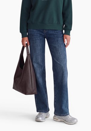 Personne portant un sweat-shirt vert foncé, un jean bleu droit, des baskets argentées, tenant un grand sac à main marron foncé dans la main gauche.
