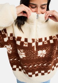 Pullover in pile a mezza zip di colore crema e marrone, con superficie texturizzata e motivi geometrici. Collo alto e vestibilità rilassata.