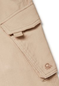 Béžová bavlněná tkanina s detaily skládací kapsy a vyšívanými akcenty, s malým výšivkovým logem poblíž spodního okraje.