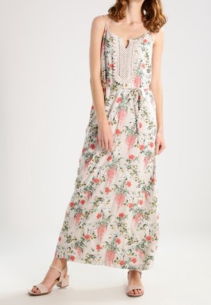 Femme portant une robe maxi sans manches à motifs floraux avec des détails en dentelle au niveau de l'encolure et des sandales beige à talons bas, debout devant un fond uni.