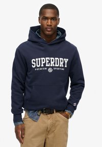 Marineblauwe hoodie met grote witte "SUPERDRY" tekst, kangoeroezak en een gestructureerde stof. Draagt over een denimshirt en beige broek.