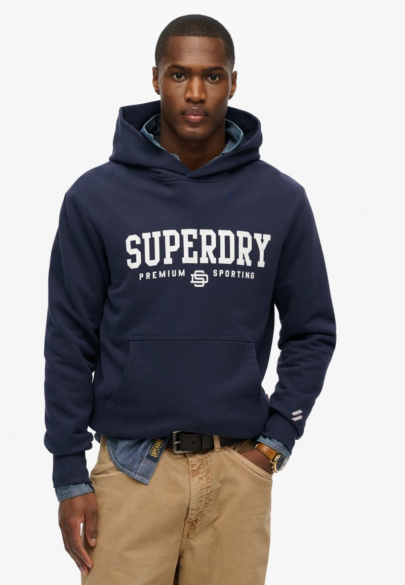 Marineblauwe hoodie met grote witte "SUPERDRY" tekst, kangoeroezak en een gestructureerde stof. Draagt over een denimshirt en beige broek.