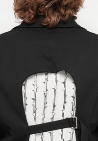 Schwarzes Kleidungsstück mit einem Ausschnitt-Design auf der Rückseite, das ein weißes Innenfutter mit einem schwarzen Dornmuster und einem schwarzen Riemen-Detail zeigt.