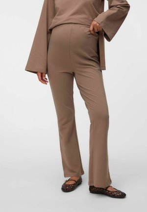 Femme portant un pantalon marron ample et un haut à manches longues assorti, debout avec une main sur la hanche, portant des ballerines tissées noires sur un sol gris.