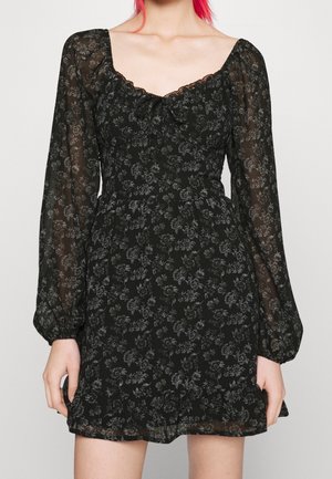 Kvinde iført en sort blomsterprint mini-kjole med gennemsigtige lange ærmer og flæsekant, stående mod en ensfarvet baggrund.
