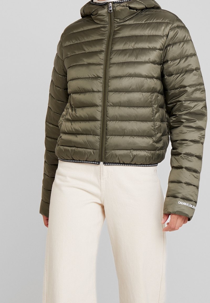 Olivgrüne Pufferjacke mit einem Reißverschluss, Kapuze und horizontalem Steppmuster. Mit markierten Akzenten am Saum und an den Ärmeln.