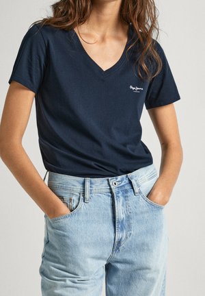 Femme portant un t-shirt bleu marine à col en V et un jean taille haute bleu clair, les mains dans les poches, sur un fond uni.