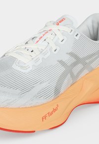 Λευκό και γκρι παπούτσι τρεξίματος ASICS με λευκά κορδόνια, πορτοκαλί ενδιάμεση σόλα με ετικέτα «FF Turbo²» και ορατό πάνω μέρος από δίχτυ.