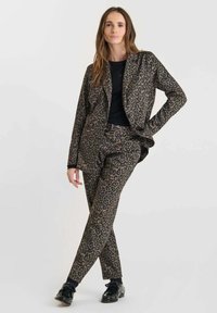 Vrouw staat met één hand op de heup, gekleed in een blazer en broek met luipaardprint, een zwart topje en zwarte schoenen, tegen een effen achtergrond.
