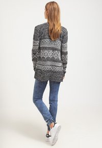 Grå mönstrad cardigan med en blandning av geometriska mönster, kombinerad med blå tapered jeans och vita sneakers. Mjuk textur, avslappnad passform.