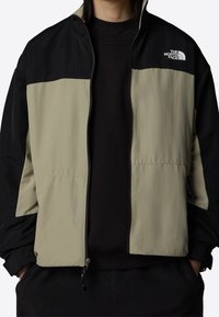 Farveblokeret jakke i beige og sort, lavet af letvægtsstof, med fuld lynlås, ståkrave og logo på brystet.