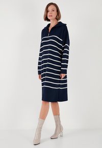 Rochie bleumarin în dungi cu guler cu fermoar, mâneci lungi și croi lejer. Asortată cu cizme bej lucioase până la gleznă cu toc pătrat transparent.